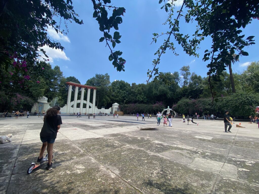 A girl stands on a scooter in a park square shaded by bougainvillea trees